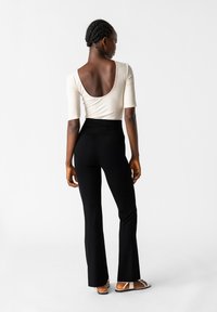 Crème ribgebreide top met een diepe ronding aan de achterkant, gecombineerd met hoge zwarte wijde broeken. Model draagt ook platte sandalen.