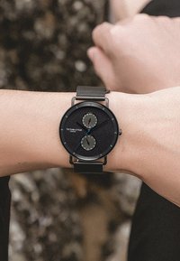 Orologio con cinturino in rete nera, quadrante rotondo con due sottodiali, sfondo nero testurizzato, dettagli in argento e un logo in basso.