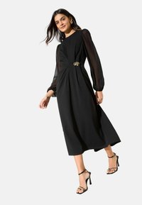 Sort langærmet kjole med tætsiddende krop, plisseret talje og en guld-accentbeslag. Modellen bærer sorte, remmede højhælede sandaler.