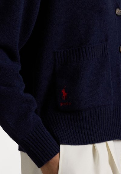 Merino villatakki tummansinisestä villasta, jossa on ribbumaiset hihansuut ja tasku, ja jossa on pieni punainen "Polo" -logo. Napit ovat näkyvissä, lisäten yksityiskohtia etupintaan.