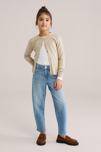 Tüdruk seisab kergelt taustal, käsi puusal, kandes beežikat kardigani, valget särki, siniseid teksapükse väikeste kaunistustega ning pruune mokasiine.