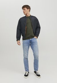 Sort bomberjakke med lynlås fortil, grøn t-shirt under, lyseblå jeans og sorte sneakers med hvid sål.