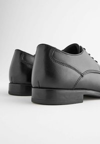 Zwarte leren schoenen met een gladde afwerking, lage hak en gestikte accenten. Het beeld toont de achterkant, waarbij de vorm en textuur worden benadrukt.