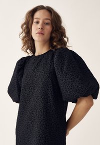 Tânără cu piele deschisă și păr castaniu creț, purtând o rochie neagră cu mâneci bufante și texturate, pe un fundal neutru.