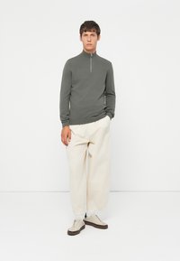 Tettsittende genser med glidelås i olivengrønn, med lange ermer. Kombinert med løstsittende kremfargede bukser og lyse joggesko med brune detaljer.