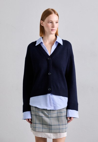 Cardigan en tricot marine à col en V profond, avec cinq boutons noirs, poignet côtelé, superposé sur une chemise bleu clair et une jupe à carreaux.