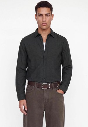 Jeune homme aux cheveux courts et bouclés, portant une chemise sombre boutonnée rentrée dans un pantalon marron avec une ceinture marron, debout sur un fond blanc.