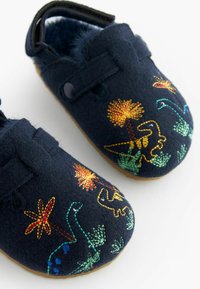 Pantoufles en feutre bleu marine foncé avec un intérieur doux, ornées d'une broderie colorée de dinosaures et de plantes sur le dessus et d'une semelle en caoutchouc beige.