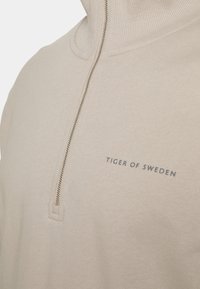 Pullover beige con colletto alto e una cerniera corta. Presenta un tessuto testurizzato e una stampa del logo in grigio scuro sul lato sinistro.