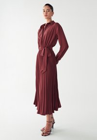 Pitkä, ruosteenvärinen mekko, jossa on kauluksellinen pääntie, napitettava etuosa, vyötärölle kuuluva vyö ja laskostettu hame. Yhdistetty mustiin remmikorkokengiin.