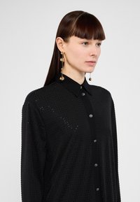 Camicia nera con bottoni, caratterizzata da un motivo testurizzato di piccoli punti. Include un colletto classico e orecchini a goccia decorativi in oro.