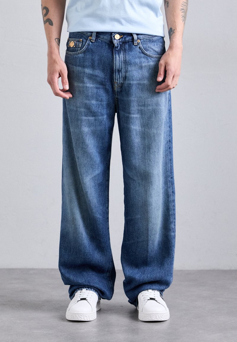 Person wearing loose-fit blue jeans and white sneakers standing on a gray floor against a plain light background.