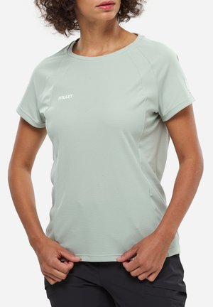 Femme ajustant un t-shirt de sport Millet vert clair à manches courtes, portant un pantalon noir, cadrée au niveau du menton sur fond blanc.