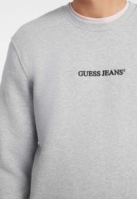 Sudadera gris de mezcla de algodón, con cuello redondo y el logo bordado en negro "GUESS JEANS" en el pecho.
