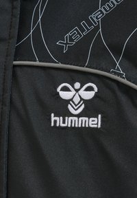 Sort jakke stof med hvid stikning, der har et fremtrædende hummel logo og abstrakte hvide linjer. Glat tekstur og moderne design.