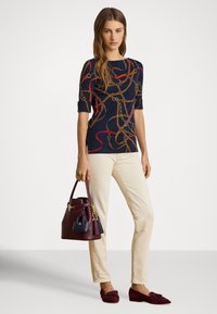 Ženska, ki nosi mornarsko modro majico z verižnim tiskom, kremne hlače, bordo loaferje ter drži ujemajočo bordo torbico z majhno mornarsko modro torbico.