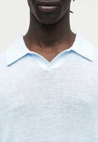 Gros plan d'une personne portant un pull en maille bleu clair avec un large col et une petite ouverture en V.