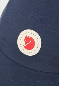Tengerészeti kerékpáros sapka texturált anyagból, kör alakú logóval, amelyen egy vörös róka dizájn látható, és a "CYCLING EQUIPMENT FJÄLLRÄVEN" felirattal.