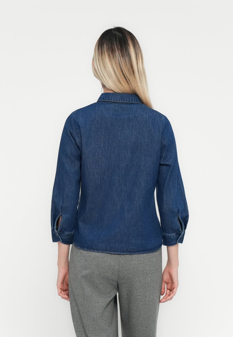 Chemise en denim bleu foncé, avec un col, des manches trois-quarts avec des détails de bouton, et une coupe décontractée. Portée avec un pantalon à motif gris.
