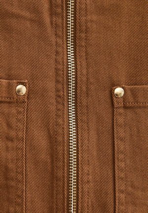 Zbliżenie na brązową lnianą kurtkę z centralnym metalowym zamkiem błyskawicznym i dwoma przednimi kieszeniami zabezpieczonymi metalowymi zatrzaskami.