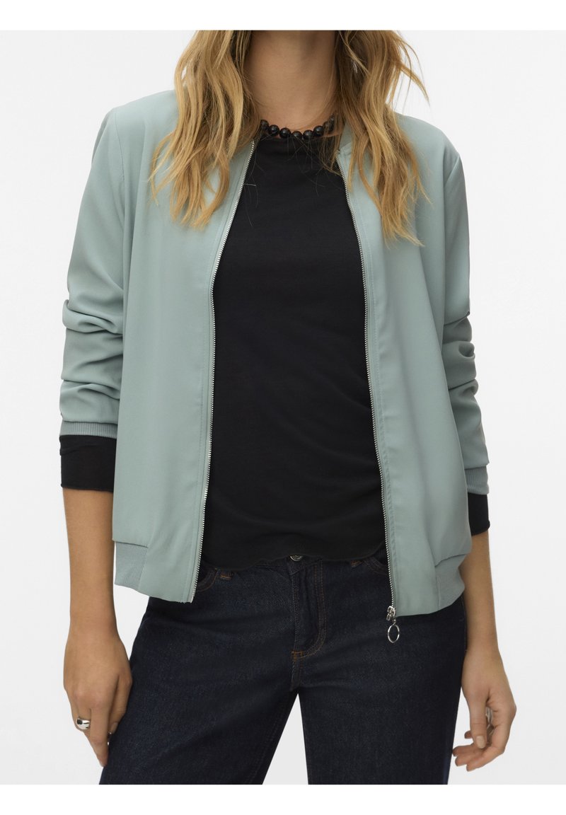 Femme portant une veste zippée vert clair sur un t-shirt noir avec un jean foncé et un collier de perles noir, debout, les bras relâchés.