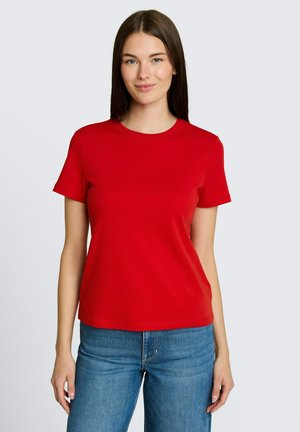 Junge Frau mit langen braunen Haaren, die ein rotes Kurzarm-T-Shirt und blaue Jeans trägt, steht vor einem einfarbigen hellen Hintergrund.
