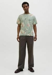 Žalias gėlėtas trumpomis rankovėmis marškinėliai su tekstūruotu raštu, akcentuotu rožinėmis ir mėlynomis detalėmis, derinami su laisvai krentančiomis tamsiai pilkomis kelnėmis ir juodais batais.