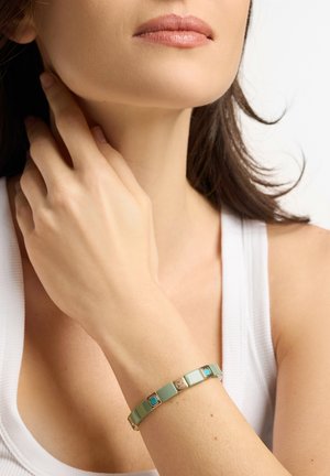 Frau trägt ein weißes Tanktop mit einem grünen und goldenen Armband am Handgelenk, Hand berührt den Hals, vor einem schlichten hellen Hintergrund.