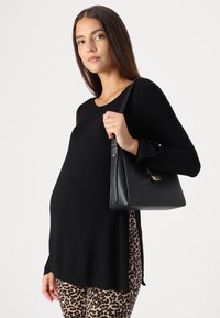 Melna Tops ar garām piedurknēm un noapaļotu apakšmalu, saskaņots ar leopardu raksta legingiem. Modelis tur struktūrizētu melnu somu ar zelta detaļām.