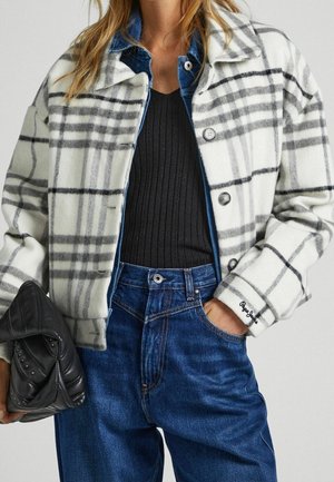 Femme portant un manteau à carreaux blanc et gris par-dessus une veste en jean et un top noir côtelé, jean bleu, tenant une pochette noire pliée.