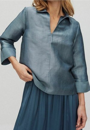 Frau trägt eine lockere, blaue, halbtransparente Bluse mit Dreiviertelärmeln und einen passenden plissierten Rock, Hand ruhend am Blusen Saum.