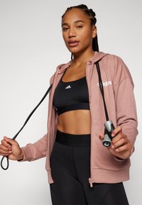Világos rózsaszín zipzáras kapucnis pulóver fekete sportmelltartó fölött, fekete leggings-szel párosítva. A modell egy fekete és szürke fogantyúval ellátott ugrókötéllel áll.