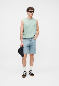 Νέος άνδρας που φοράει αμάνικο πουκάμισο σε μέντα πράσινο χρώμα, ανοιχτόχρωμα τζιν σορτς, μαύρα αθλητικά παπούτσια, λευκές κάλτσες, γυαλιά ηλίου και κρατάει μια μαύρη ριγέ καπέλο.