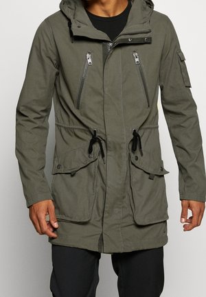 Homme portant une veste à capuche vert olive avec deux fermetures éclair poitrine, taille à cordon, poches rabat avec boutons-pression et une poche sur la manche.