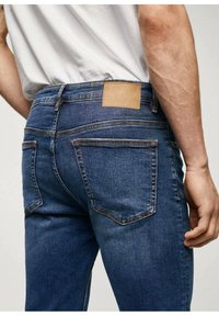Tmavě modré džíny s úzkým střihem, s koženou nášivkou na zadním pásku a kontrastním prošíváním na kapsách.