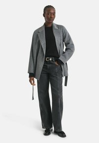 Grau melierte Wolljacke mit Reverskragen, getragen über einem schwarzen Kabelstrickpullover. Kombiniert mit hochtaillierten schwarzen Jeans. Schwarze Schuhe.