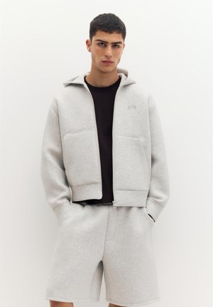 Giovane uomo con felpa leggera grigio chiaro con zip e pantaloncini abbinati sopra una maglietta nera, in piedi davanti a uno sfondo bianco semplice.