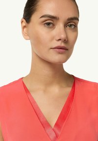 Zbliżenie na kobietę z zielonymi oczami i gładką skórą, ubraną w koralowy top w serek, lekko zwróconą w prawo na jasnym tle.