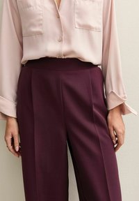 Lichtroze blouse met knoopdetails en twee borstzakken, gecombineerd met hooggetailleerde, diep bordeauxrode plooibroek. Gladde textuur.