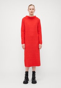 Abito lavorato a maglia rosso con scollo a cowl, maniche lunghe e silhouette dritta. Abbinato a stivali alla caviglia neri e robusti con fibbie.