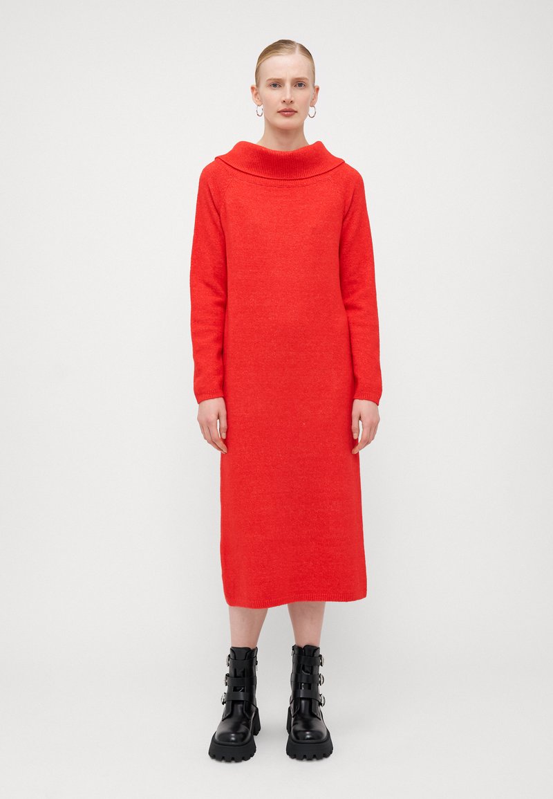Abito lavorato a maglia rosso con scollo a cowl, maniche lunghe e silhouette dritta. Abbinato a stivali alla caviglia neri e robusti con fibbie.