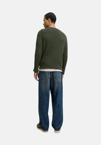 Mann, der sich abwendet und einen olivgrünen Kabelstrickpullover, lockere blaue Jeans, beigen Sneakers und ein weißes Hemd, das unter dem Pullover hervorblitzt, trägt.