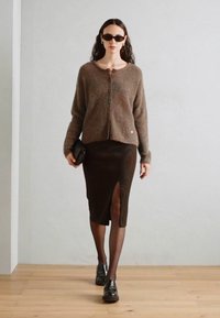 Cardigan en tricot marron avec fermeture à boutons, associé à une jupe sombre arrivant au genou avec une fente sur le côté. Le modèle porte des lunettes de soleil et des chaussures noires.