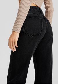 Mujer vistiendo jeans de mezclilla negros de talle alto y un top corto beige de manga larga acanalada, vista desde atrás con la mano apoyada a un lado.