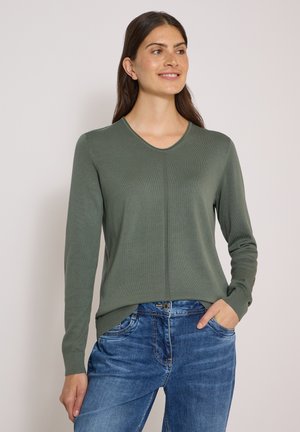 Femme aux longs cheveux portant un pull vert à manches longues et un jean bleu, souriante et regardant sur le côté avec une main dans la poche.