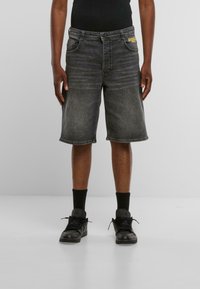 Shorts de mezclilla gris oscuro con un corte recto, ligero desgaste, cinco bolsillos y un acento de logo amarillos en la cinturilla, combinados con calzado negro.