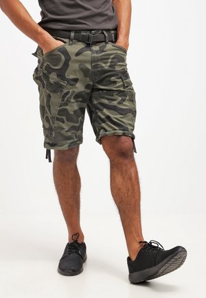 Mężczyzna w podwiniętych moro spodenkach typu cargo, czarnym pasku, szarej koszulce i czarnych trampkach, z rękami w kieszeniach, na białym tle.