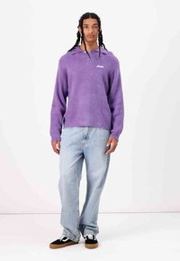 Pulover din flece violet cu guler și logo, combinat cu blugi decolorati de culoare albastră deschis și adidași negri cu accente albe.