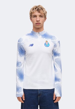 Jeune homme aux cheveux blonds courts portant un maillot de sport blanc et bleu à manches longues avec une demi-fermeture éclair et des logos d'équipe, debout devant un fond uni.