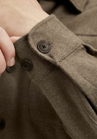 Puño de camisa de mezcla de lana marrón con cierre de botón y una textura suave, con dos botones oscuros y un borde de puño doblado de manera ordenada.